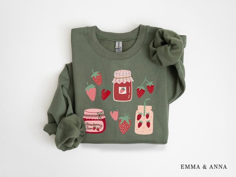 May include: A green sweatshirt with a strawberry jam and strawberry smoothie design. The design features jars of strawberry jam and a glass of strawberry smoothie with a straw, all surrounded by strawberries.