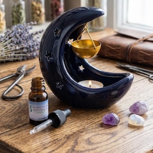 May include: A navy blue ceramic moon-shaped essential oil diffuser with star cutouts and a gold-coloured bowl. A lit candle sits inside the moon. An essential oil bottle and amethyst crystals are nearby.