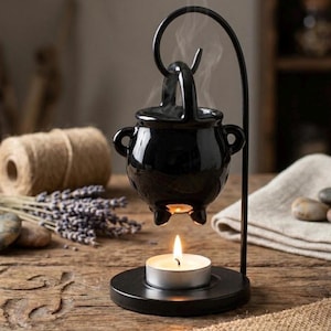 May include: A black ceramic cauldron-shaped wax warmer suspended from a curved metal stand. The piece features a small handle on top and two side handles, with a lit tea light candle placed on the circular base directly beneath the vessel.