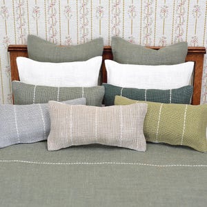 May include: A collection of miniature pillows in various shades of green, white, and beige. The pillows have a rectangular shape with vertical white stripe detailing. The pillows are arranged on a green surface, suggesting a bed setting.