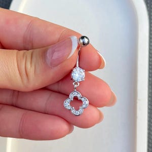 May include: A silver-toned belly button ring featuring a curved barbell with a round top ball, a central round clear stone, and a dangling four-leaf clover charm encrusted with small clear stones along its metallic border and center.