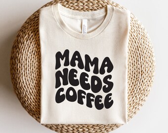 Pode incluir: Uma camiseta branca com o texto "MAMA NEEDS COFFEE" em letras pretas estilizadas.