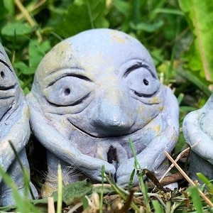 Könnte beinhalten: Drei skurrile, grau-blaue Keramikfiguren mit ausdrucksstarken Gesichtern und verschränkten Armen sitzen im grünen Gras. Die Skulpturen haben große, ovale Augen und einen leicht traurigen Ausdruck, was ein einzigartiges Dekorationsstück ergibt.