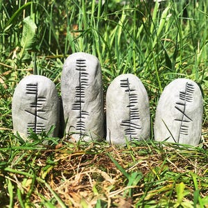 May include: Four grey stone rune stones with carved black symbols, arranged in a row and set in green grass. The stones are oval-shaped and vary slightly in height. The symbols are vertical lines with horizontal dashes.
