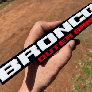 May include: A black, white, and red plastic emblem with the words "BRONCO OUTER BANKS" in raised lettering.