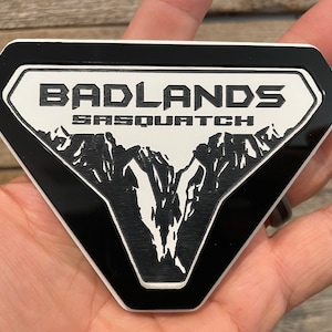 May include: Black and white emblem with a diamond shape and a black border. The emblem features the text "BADLANDS SASQUATCH" and a graphic of a mountain range.