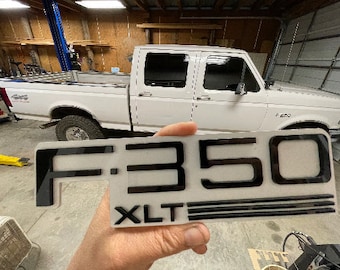 May include: Black and white plastic emblem with the text "F-350 XLT" in a bold font. The emblem has a raised, three-dimensional design.