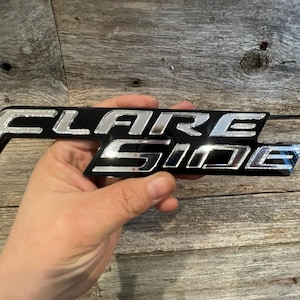 May include: Black and silver "FLARE SIDE" emblem. The emblem has a black base with the words "FLARE SIDE" in a shiny silver finish. The emblem is held in a hand.