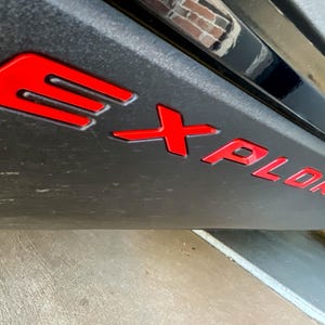 May include: Close-up of a black vehicle panel with the word "EXPLORER" in bright red, stylized lettering. The "E" is a distinctive, three-bar design. The panel has a textured, matte finish.