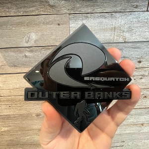 May include: Black diamond-shaped car emblem with a gray wave design and the text "Sasquatch Outer Banks".