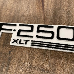 May include: White and black plastic emblem with the text "F-250 XLT" in black lettering. The emblem has three black stripes below the text.