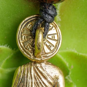 May include: Close-up of a gold-colored pendant with a circular design and a figure. The pendant is attached to a black cord. The background is a green plant.