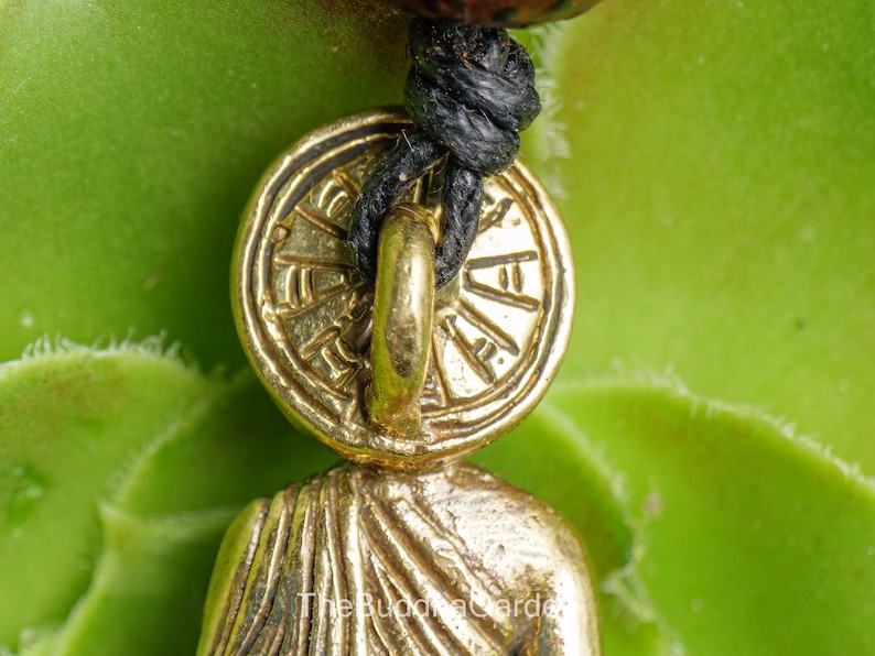 May include: Close-up of a gold-colored pendant with a circular design and a figure. The pendant is attached to a black cord. The background is a green plant.