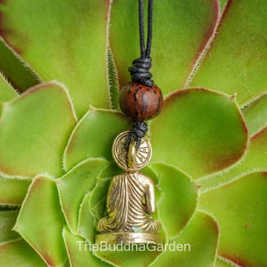 May include: A gold-colored Buddha pendant necklace with a seated figure, a round coin-like element, and a dark brown bead. The necklace is suspended from a black cord against a backdrop of green succulent leaves. The text "TheBuddhaGarden" is at the bottom.