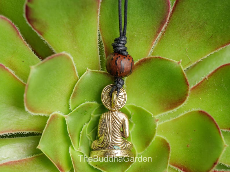 May include: A gold-colored Buddha pendant necklace with a seated figure, a round coin-like element, and a dark brown bead. The necklace is suspended from a black cord against a backdrop of green succulent leaves. The text "TheBuddhaGarden" is at the bottom.