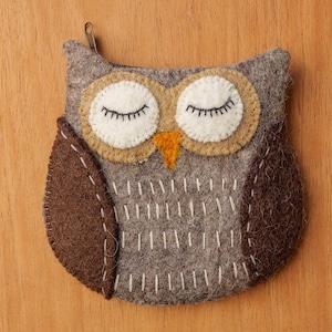 May include: A felt owl-shaped coin purse with a zipper closure. The purse features a gray body, brown wings, and white and tan eyes with black eyelashes. The owl has an orange beak and is decorated with white stitching.