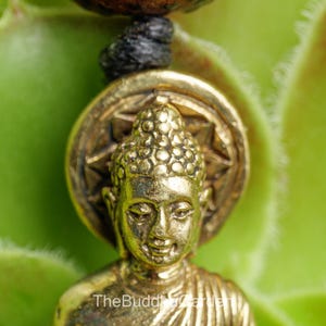 May include: Close-up of a gold-colored Buddha pendant. The pendant features a detailed Buddha figure with a textured head and a starburst design behind it. The background is a blurred green.