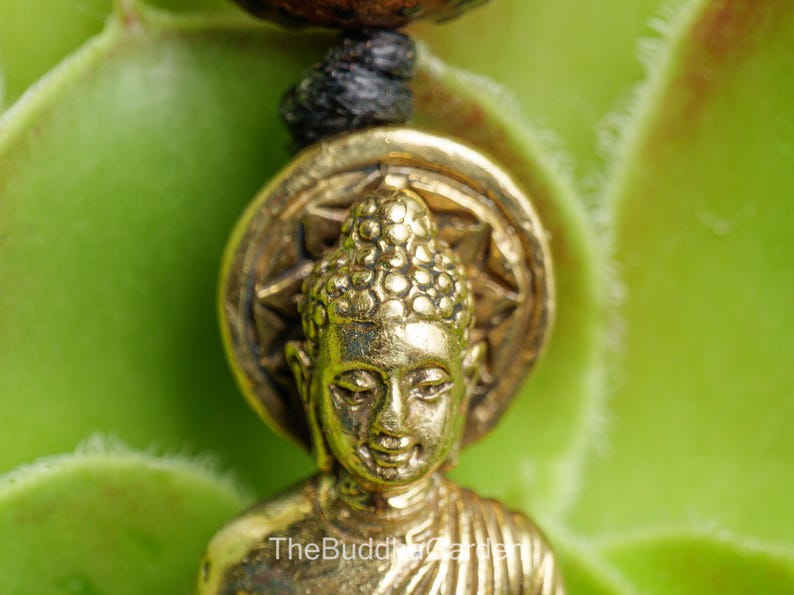 May include: Close-up of a gold-colored Buddha pendant. The pendant features a detailed Buddha figure with a textured head and a starburst design behind it. The background is a blurred green.