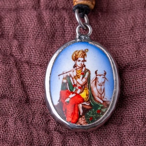 May include: A silver pendant featuring a colourful image of a Hindu deity playing a flute and riding a cow. The pendant is displayed on a brown fabric background.