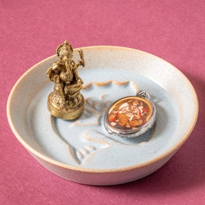 May include: A small, gold-colored statue of Ganesha, the Hindu god of wisdom and prosperity, sits on a light blue ceramic dish. A silver pendant with a colorful image of Ganesha is also on the dish.