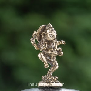 May include: A small, gold-colored brass statue of Ganesha, the Hindu god of beginnings, prosperity, and good fortune. The statue depicts Ganesha in a dancing pose, with his trunk raised and his hands in a mudra gesture. The statue is set against a green background.