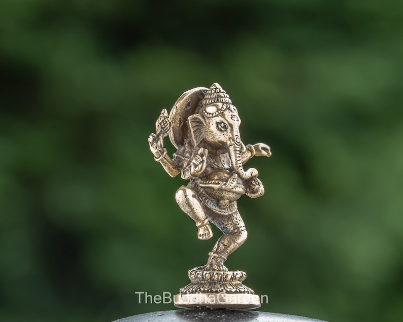 May include: A small, gold-colored brass statue of Ganesha, the Hindu god of beginnings, prosperity, and good fortune. The statue depicts Ganesha in a dancing pose, with his trunk raised and his hands in a mudra gesture. The statue is set against a green background.