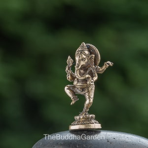 May include: A small, gold-colored metal statue of Ganesha, the Hindu god of beginnings, wisdom, and prosperity. The statue depicts Ganesha in a dancing pose with his trunk raised and his hands in a mudra gesture. The statue is set on a small, circular base.