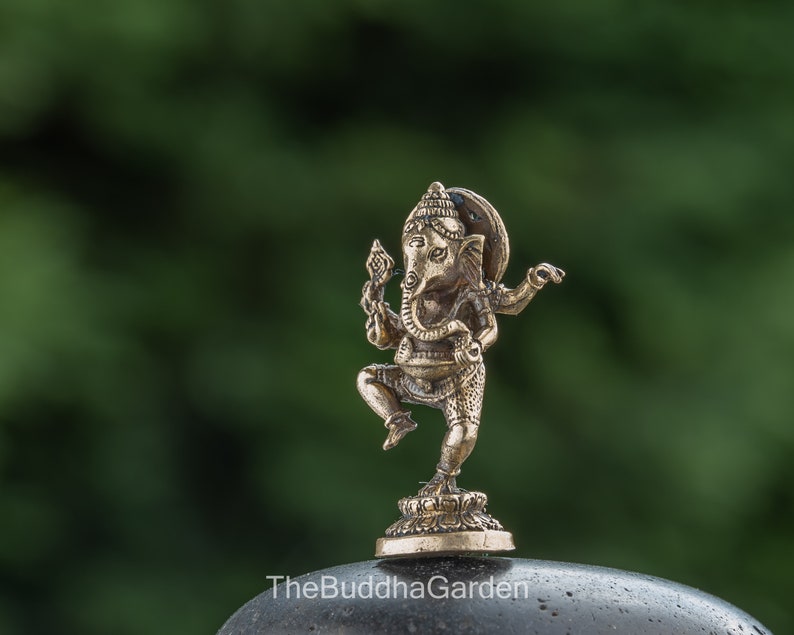 May include: A small, gold-colored metal statue of Ganesha, the Hindu god of beginnings, wisdom, and prosperity. The statue depicts Ganesha in a dancing pose with his trunk raised and his hands in a mudra gesture. The statue is set on a small, circular base.