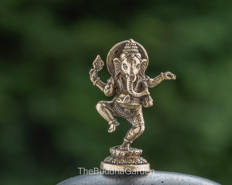 May include: A small, gold-colored brass statue of the Hindu deity Ganesha. The statue depicts Ganesha in a dancing pose, with his trunk raised and his hands in a gesture of blessing. The statue is set on a small, circular base.