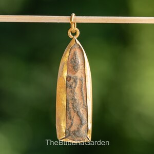 May include: A gold-plated pendant featuring a brown and gray Buddha image. The pendant is shaped like a teardrop and has a loop at the top for attaching a chain.