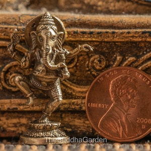 May include: A small, gold-colored metal statue of the Hindu deity Ganesha. The statue is in a dancing pose, with one leg raised and the other bent. Ganesha is depicted with an elephant head, four arms, and a large belly. The statue is about the size of a US penny.