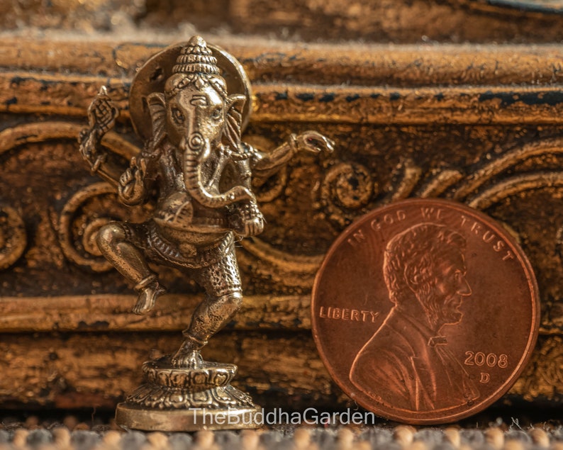 May include: A small, gold-colored metal statue of the Hindu deity Ganesha. The statue is in a dancing pose, with one leg raised and the other bent. Ganesha is depicted with an elephant head, four arms, and a large belly. The statue is about the size of a US penny.