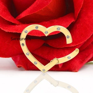 May include: A gold heart-shaped charm with clear sparkling stones. The charm is sitting on a white surface with a red rose in the background.