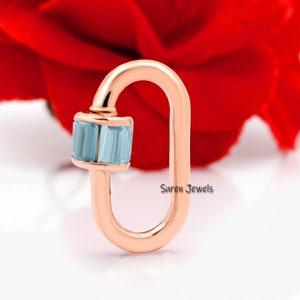 May include: Rose gold charm featuring a light blue gemstone. The charm is shaped like a sideways oval with a small loop at the top. The charm is displayed against a white background.