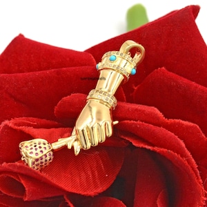 Op de afbeelding: Een gouden hanger met een hand die een roos vasthoudt. De hand is versierd met een kroon en turquoise stenen. De roos is rood en heeft een groene steel.