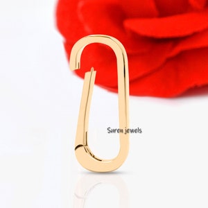 May include: A gold-colored metal clasp with a rounded oval shape. The clasp is designed to attach to a chain or other jewelry piece. The clasp is shown against a white background with a red rose in the background.