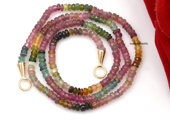 May include: A multi-colored gemstone necklace with gold clasps. The necklace is made of small, round beads in shades of pink, green, brown, and black. The necklace is lying on a white surface with a red rose in the background.