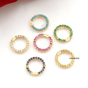 Op de afbeelding: Zes gouden ringen met een enkele rij gekleurde edelstenen. De ringen zijn in een halve cirkel op een witte achtergrond gerangschikt. De edelstenen zijn turquoise, roze, groen, blauw, geel en wit.