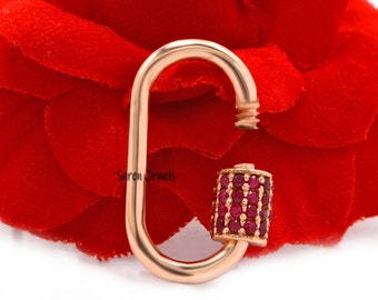 May include: Rose gold charm featuring a red ruby stone accent. The charm is shaped like a carabiner clip and has a small, cylindrical ruby stone on the bottom.