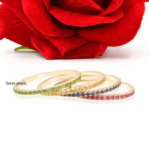 May include: Four gold stacking rings with a row of tiny gemstones. The rings are stacked in order of color: green, yellow, blue, and red.