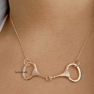 May include: A gold-toned necklace with a horse bit pendant. The pendant is made of two connected rings with a small bar connecting them.