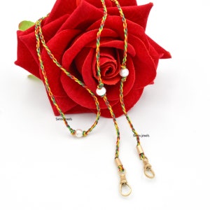 Puede incluir: Un collar de cordón trenzado rojo, verde y amarillo con tres cuentas de perla blanca y cierres dorados. "Surenu Jewels" está impreso en el cordón.