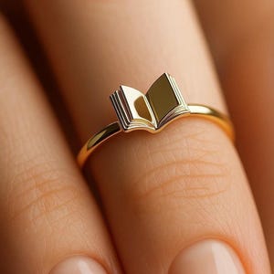Könnte beinhalten: Ein goldener Ring mit einem Buchdesign. Das Buch ist geöffnet und die Seiten sind sichtbar. Der Ring befindet sich an einem Finger.