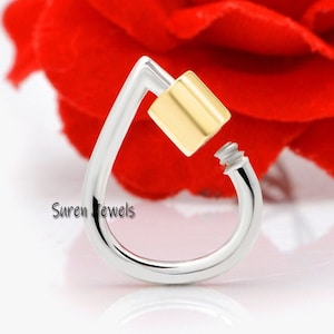 Puede incluir: Una joya plateada en forma de lágrima con un acento cuadrado de color dorado. La pieza tiene un detalle en forma de tornillo en el extremo y el texto "Suren jewels" es visible. El fondo es rojo borroso.