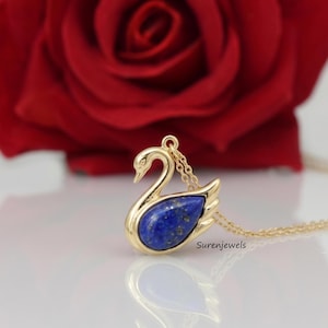 May include: A gold-toned swan pendant necklace featuring a deep blue lapis lazuli stone set in the centre of the swan's body. The pendant is attached to a delicate gold-toned chain. The text Surenjewels is printed near the base of the pendant.