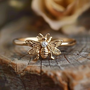May include: A gold ring featuring a detailed bee design with a small clear stone at its centre.