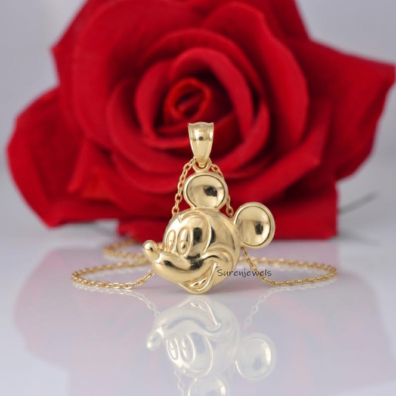 May include: A polished gold pendant shaped like the head of Mickey Mouse, suspended from a delicate gold chain. The pendant features a smooth, reflective surface with the engraved text Surenjewels visible on the lower right side of the design.