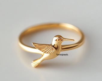 Könnte beinhalten: Ein goldfarbener Ring mit einem detaillierten Kolibri-Design. Der Vogel ist im Flug dargestellt, mit ausgebreiteten Flügeln und einem langen Schnabel. Der Ring hat eine glatte, polierte Oberfläche, und der Kolibri ist das Hauptmerkmal des Schmucks.