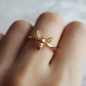 May include: A gold-coloured ring with a detailed bee design. The bee has textured wings and body, with the ring band extending around the finger. The ring is a close-up shot, highlighting the intricate details of the jewellery.