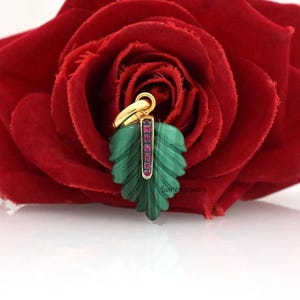 May include: A gold pendant shaped like a carved green malachite leaf with a central vertical bar set with small pink gemstones. The pendant features a gold bail at the top and is positioned against a large, deep red rose blossom.